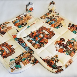 May include: Two quilted pot holders with a fall theme. The pot holders are cream colored with a patchwork design and feature a red truck, pumpkins, scarecrows, and sunflowers.
