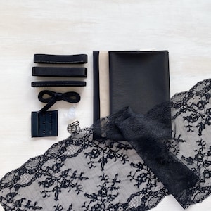 May include: An assortment of black sewing supplies, including fabric, lace trim, elastic, and a hook-and-eye closure. The lace features a floral pattern. The items are arranged on a light-colored surface, suggesting a craft or DIY project.