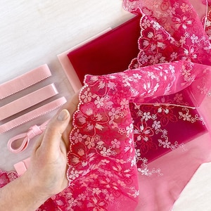 May include: An assortment of pink ribbons, lace, and fabric. The image features various shades of pink, including a deep magenta, light pink, and a sheer, floral-patterned lace. The items are arranged on a white surface, showcasing the textures and colors.