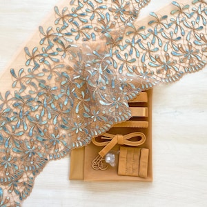 May include: A roll of beige lace with a floral design in brown and blue. The lace is delicate and detailed, with a scalloped edge. Also included are beige bra straps, a hook and eye closure, and a bra strap extender.