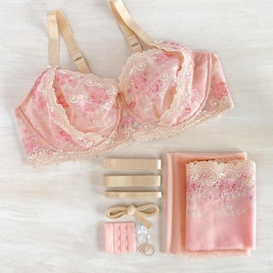 May include: A pink bra with floral embroidery and beige straps, lace trim, and matching accessories. The set includes extra straps, a bow, hook-and-eye closures, and fabric. The bra is a delicate, feminine design.
