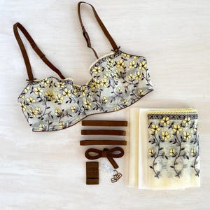 May include: A lingerie set featuring a bra with yellow floral embroidery on sheer fabric, brown straps, and matching accessories. The set includes extra straps, a bow, and a fabric panel with similar floral detailing.