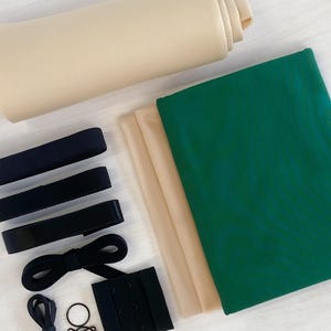 May include: A set of sewing supplies for a DIY project, including green and beige fabric, black elastic, and a bra clasp.
