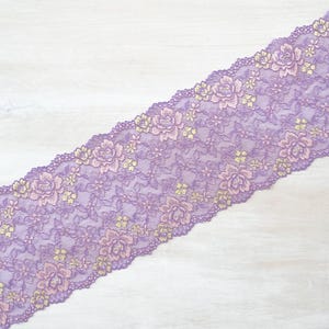 May include: Purple lace trim with a floral pattern in a lighter shade of purple and gold. The lace is on a white wooden surface.