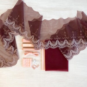 May include: A collection of sewing and lingerie-making supplies. Includes burgundy lace fabric with a scalloped edge, pink ribbons, bra straps, and other accessories. The items are arranged on a light wooden surface.