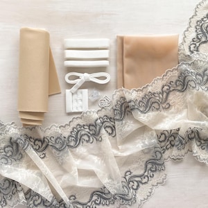 May include: A collection of lingerie-making supplies, including beige fabric rolls, white elastic, a hook and eye closure, and a delicate lace trim with a gray floral design. The items are arranged on a light wooden surface.