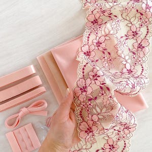 May include: A collection of pink lingerie-making supplies. Includes a floral lace trim, fabric, elastic, hook and eye closures, and a bow. The lace features a cream background with pink floral designs. The items are arranged on a white surface.