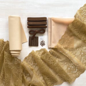 May include: An assortment of lingerie-making supplies, including a roll of beige fabric, brown elastic, a hook and eye closure, and a length of gold lace trim. The items are arranged on a white surface, showcasing the materials for crafting intimate apparel.