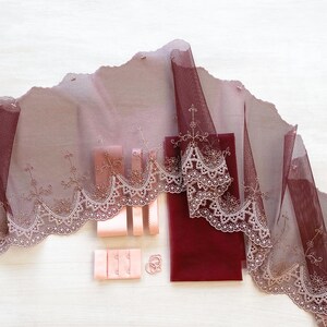 May include: A collection of burgundy and blush-colored fabric and sewing notions. The image features a length of embroidered lace trim, satin ribbon, bra closures, and a small piece of solid fabric, all arranged on a white surface.