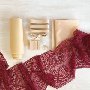 May include: A collection of lingerie-making supplies, including a roll of beige fabric, beige elastic, a hook and eye closure, and a length of burgundy lace with a geometric pattern. The items are arranged on a light-colored surface.