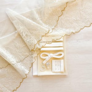 May include: Ivory-colored lace fabric with floral embroidery and a scalloped gold trim. A kit containing white ribbons, a bow, and other accessories is also visible. The items are arranged on a light-colored surface.