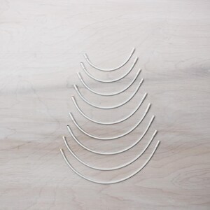 May include: A set of nine silver-colored, graduated, circular sewing needles. The needles are arranged in a pyramid shape on a light wood surface.