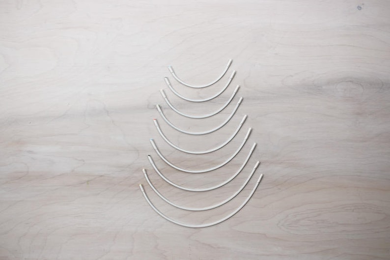 May include: A set of nine silver-colored, graduated, circular sewing needles. The needles are arranged in a pyramid shape on a light wood surface.
