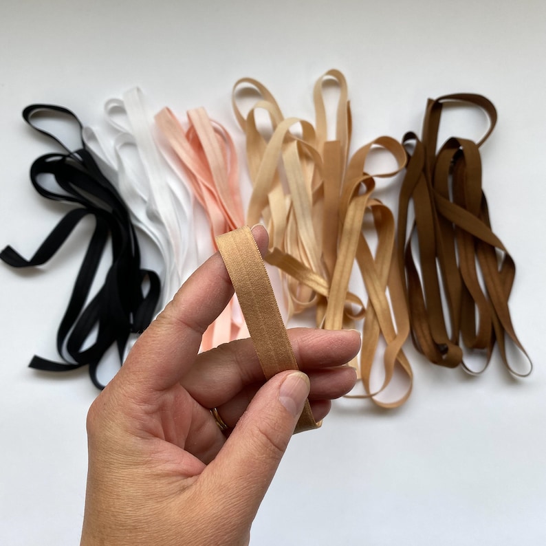 May include: Assortment of elastic ribbon in various colors, including black, white, beige, peach, and brown. The ribbon is approximately 1/2 inch wide.