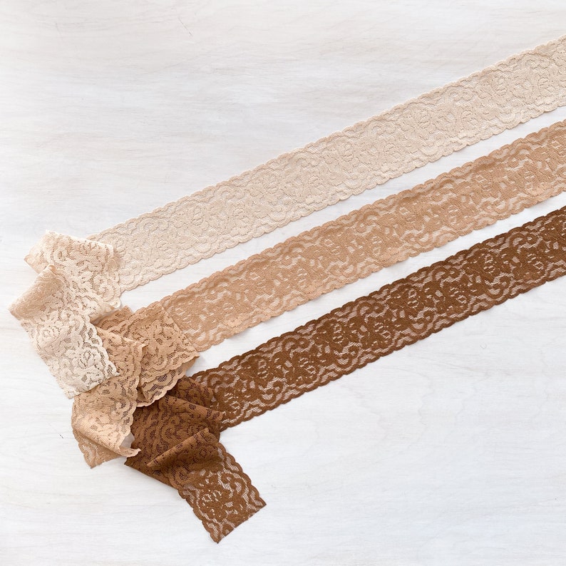 May include: Three different lace trims in beige, brown, and dark brown colors. The lace trims are arranged in a diagonal pattern on a white surface.