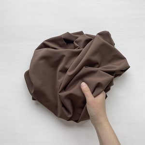 May include: A brown fabric swatch, crumpled and held in a hand. The fabric is smooth and has a slight sheen.