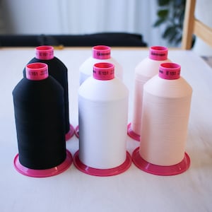 Peut inclure: Quatre bobines de fil Gutermann en noir, blanc et rose clair. Chaque bobine porte une étiquette rose avec le texte "Gutermann E 151 100m 100% Polyester" imprimé dessus.