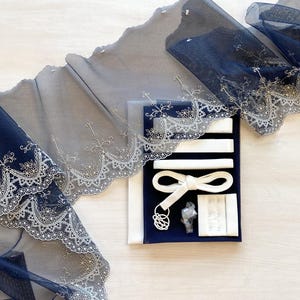 May include: Navy blue lace trim with a scalloped edge and floral embroidery. A kit includes white ribbon, a bow, a hook and eye closure, and other sewing notions. The items are arranged on a light wood surface.