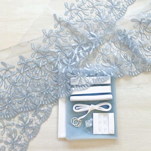 May include: A light blue floral lace fabric with a scalloped edge, alongside a sewing kit containing white elastic, a hook and eye closure, and other notions. The lace features a delicate embroidered flower pattern.