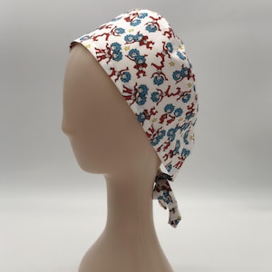 May include: White surgical scrub cap featuring a repeating pattern of red and blue cartoon characters and yellow stars. The cap is tied at the back and is displayed on a mannequin head.