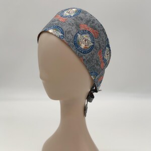 May include: Gray surgical scrub cap with a repeating pattern of circular emblems in red, white, and blue. The emblems feature an eagle and the text "Aerosmith Force One". The cap is tied at the back.
