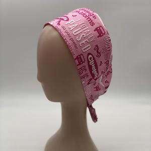 May include: A pink headscarf with a repeating pattern of text and small vehicle illustrations. The text includes phrases like "I totally paused" and "Clueless". The head covering is tied at the back.