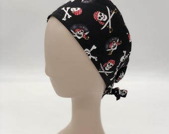 May include: A black scrub cap featuring a repeating pattern of white skulls wearing red and white polka-dot bandanas, accompanied by crossed swords. The cap has a tie-back closure at the nape of the neck and a contoured fit for the head.
