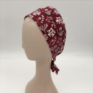 May include: A maroon and white patterned scrub cap featuring the text "Texas A&M" and various Aggie-themed graphics. The cap is tied at the back and is displayed on a mannequin head.