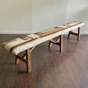 May include: A long wooden bench with a white and brown patterned cushion. The bench has a rustic, farmhouse style and is perfect for adding a touch of warmth to any room.