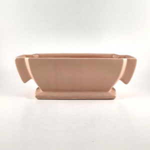 May include: A pale pink ceramic planter with a rectangular shape and two angled wings on each side. The planter has a smooth, glossy finish.