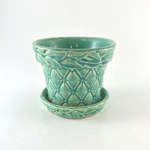 May include: A teal ceramic plant pot with a matching saucer. The exterior features a quilted diamond pattern with a raised, braided leaf design around the top rim. The glaze has a glossy finish, highlighting the textured details of the vessel.