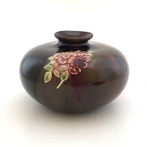 May include: A small, round, dark brown ceramic vase with a floral design. The vase has a glossy finish and features a cluster of pink and green flowers.
