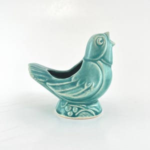 May include: A teal ceramic bird-shaped planter. The bird has a detailed design with visible feathers and a small opening for planting. The planter sits on a round base with a decorative leaf and berry pattern.
