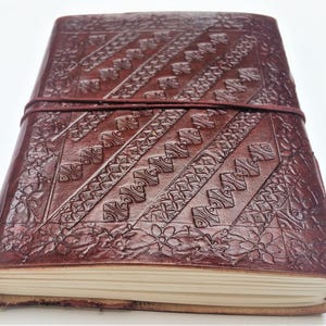 May include: A handcrafted, dark brown leather journal with intricate embossed patterns. The cover features diagonal geometric designs and floral accents, secured by a matching leather strap. The journal's pages are visible, suggesting a blank notebook.