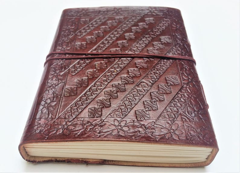 May include: A handcrafted, dark brown leather journal with intricate embossed patterns. The cover features diagonal geometric designs and floral accents, secured by a matching leather strap. The journal's pages are visible, suggesting a blank notebook.