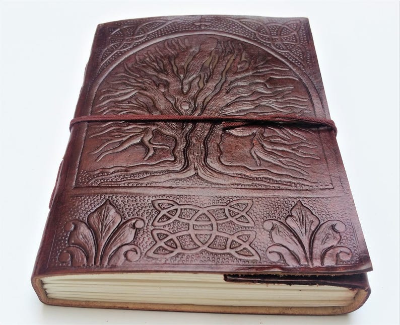May include: A brown leather journal with an embossed tree design on the cover. The journal is secured with a burgundy elastic band. The cover also features Celtic knot and leaf designs. The pages are cream-colored.