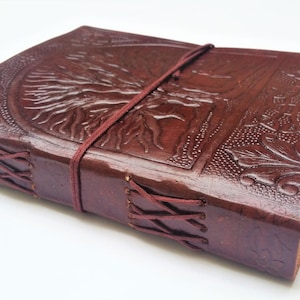 May include: A brown leather journal with an embossed tree of life design on the cover. The journal is secured with a burgundy elastic band and features stitched edges. The overall aesthetic is rustic and handcrafted.
