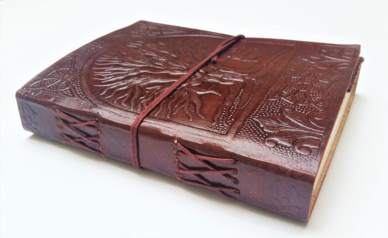 May include: A brown leather journal with an embossed tree of life design on the cover. The journal is secured with a burgundy elastic band and features stitched edges. The overall aesthetic is rustic and handcrafted.