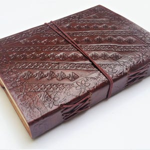 May include: A dark brown leather journal with intricate embossed patterns and a matching elastic closure. The cover features geometric and floral designs. The journal is bound with visible stitching and has a rustic, handcrafted appearance.
