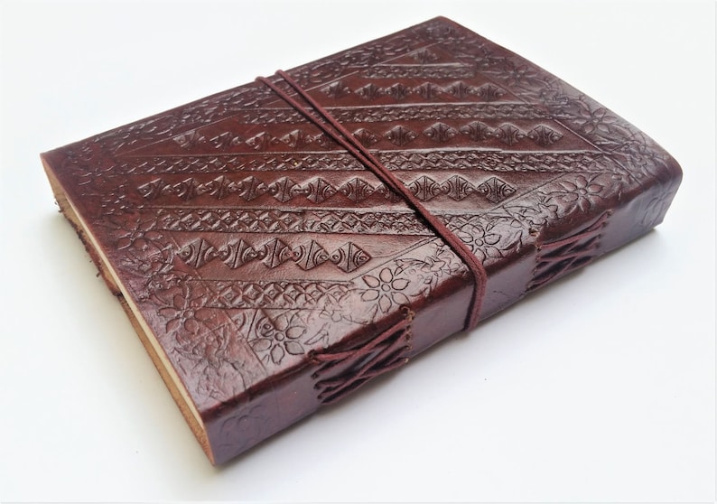 May include: A dark brown leather journal with intricate embossed patterns and a matching elastic closure. The cover features geometric and floral designs. The journal is bound with visible stitching and has a rustic, handcrafted appearance.