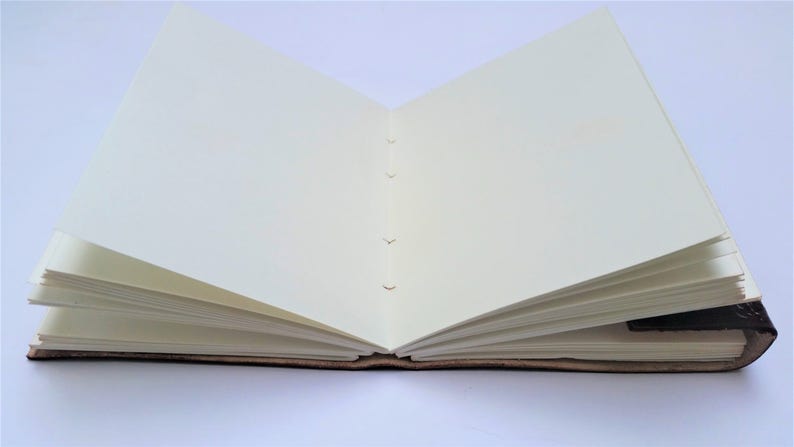 May include: An open, handmade journal with cream-colored pages. The journal's spine is visible, showing the binding stitches. The cover appears to be a dark brown, possibly leather, with a textured finish. The pages are blank and ready for writing or sketching.