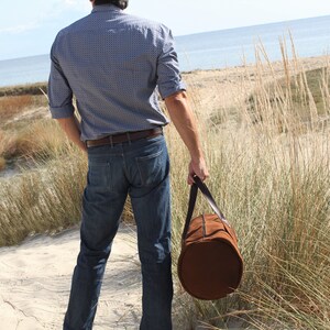 A male model holds a round, tobacco-colored, true Italian leather handcrafted travel bag.