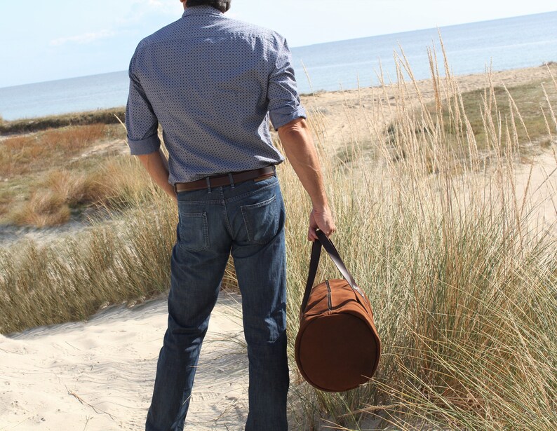 A male model holds a round, tobacco-colored, true Italian leather handcrafted travel bag.