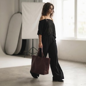 May include: A black off-the-shoulder top with gold chain straps paired with matching black wide-leg trousers, accompanied by a large, structured dark burgundy leather tote bag with dual handles, creating a coordinated ensemble.