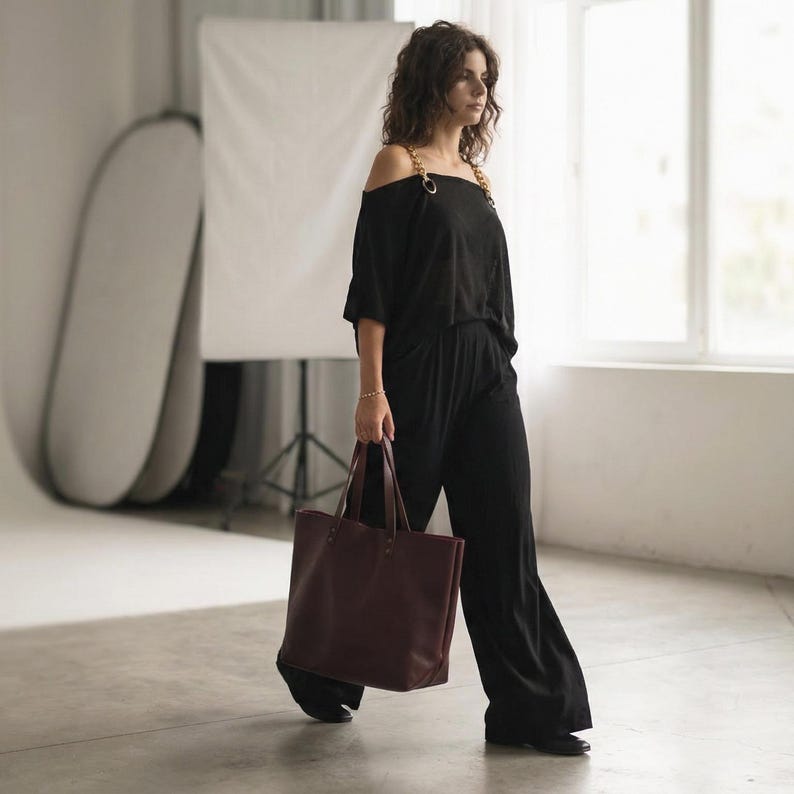 May include: A black off-the-shoulder top with gold chain straps paired with matching black wide-leg trousers, accompanied by a large, structured dark burgundy leather tote bag with dual handles, creating a coordinated ensemble.