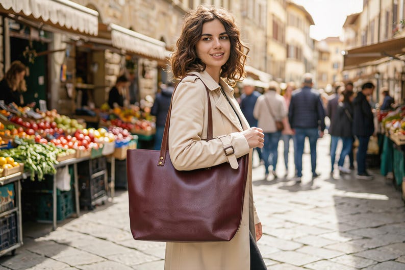 May include: A deep burgundy leather tote bag with two thin shoulder straps and gold-toned metal hardware attachments. The bag features a wide, open-top rectangular design with a smooth, solid-colored finish and clean, structured stitching along the sides.
