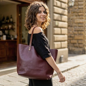 May include: A large, deep burgundy leather tote bag with two long shoulder straps attached by metallic rivets. The bag features a smooth, matte finish and a rectangular structure, providing a spacious interior compartment for carrying daily essentials.
