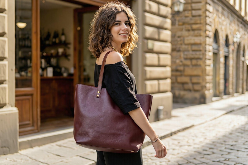 May include: A large, deep burgundy leather tote bag with two long shoulder straps attached by metallic rivets. The bag features a smooth, matte finish and a rectangular structure, providing a spacious interior compartment for carrying daily essentials.
