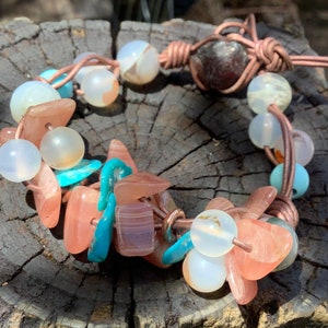 May include: A handmade bracelet featuring a mix of natural stones and beads. The bracelet includes peach, turquoise, and white beads, along with a dark red stone. The stones are strung on brown leather cord.