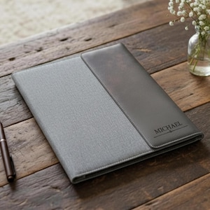 May include: A grey and dark grey personalised portfolio with the name "MICHAEL" embossed on the front. The portfolio is made of a textured material and is on a wooden surface. A pen is next to the portfolio.